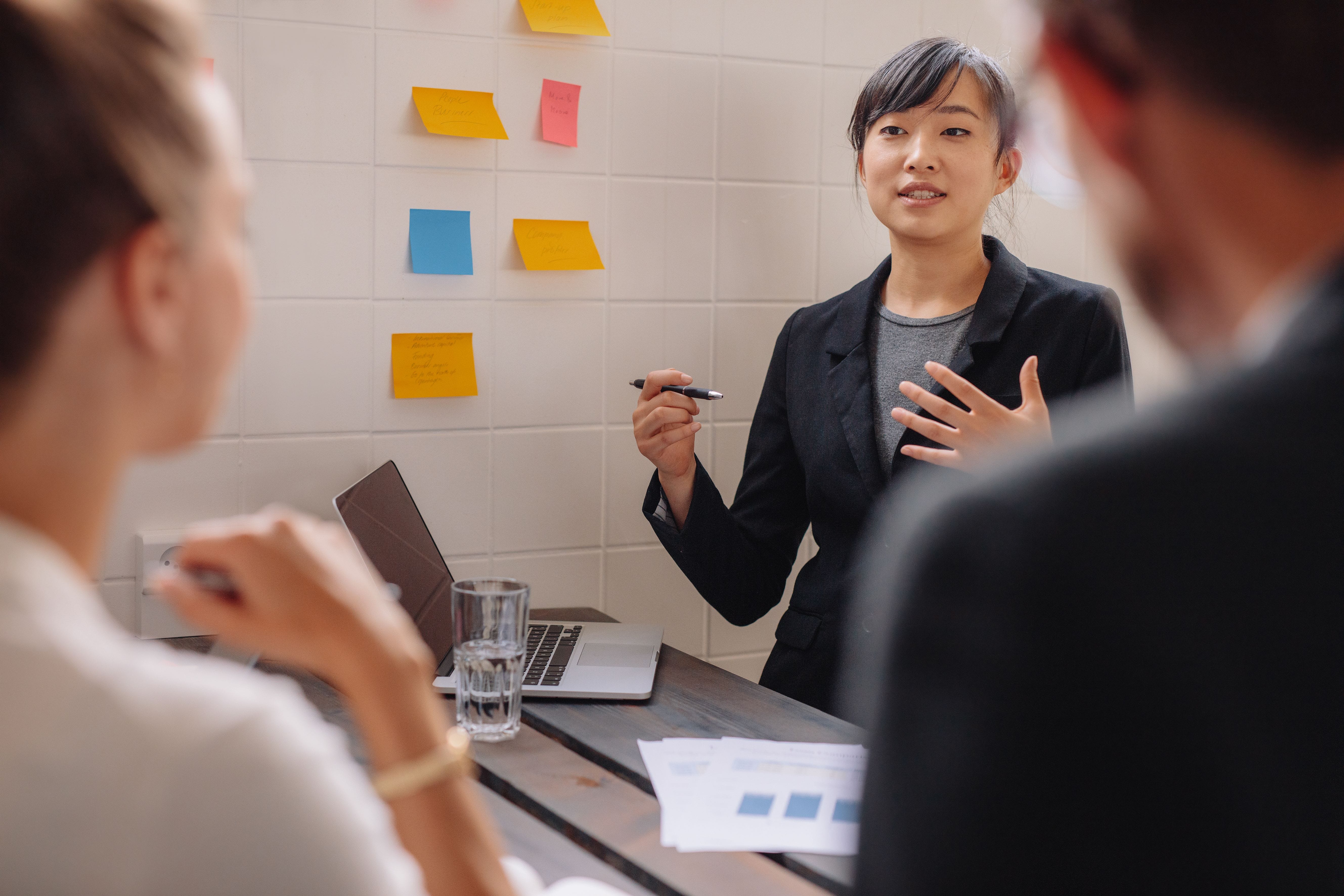Team meeting in progress, with a presenter speaking beside a wall of sticky notes while colleagues listen around a conference table.